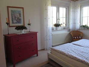 Une chambre avec une commode rouge, un lit recouvert d’une couvre-lit à motifs floraux, un fauteuil en osier et une fenêtre avec des rideaux.