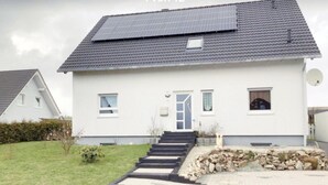 Una casa a un piano con tetto spiovente, pannelli solari, un portone d'ingresso e un piccolo giardino antistante con un muro in pietra e delle scale che conducono all'entrata.