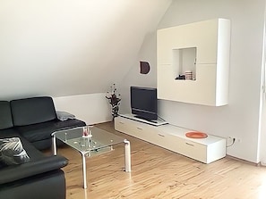 Un salon moderne avec un canapé noir, une table basse en verre, un téléviseur à écran plat posé sur un meuble de télévision blanc et une petite plante.