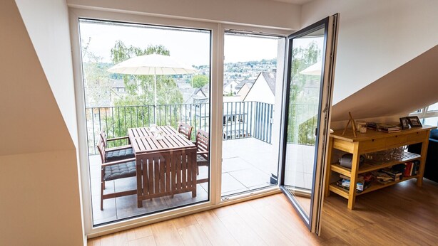 A room with a wooden table and chairs, a bookshelf, and a sliding glass door leading to an outdoor balcony with a view of buildings and trees. - Apartment 'Ferienwohnung Weitblick Koblenz' with Balcony and Wi-Fi (Koblenz)
