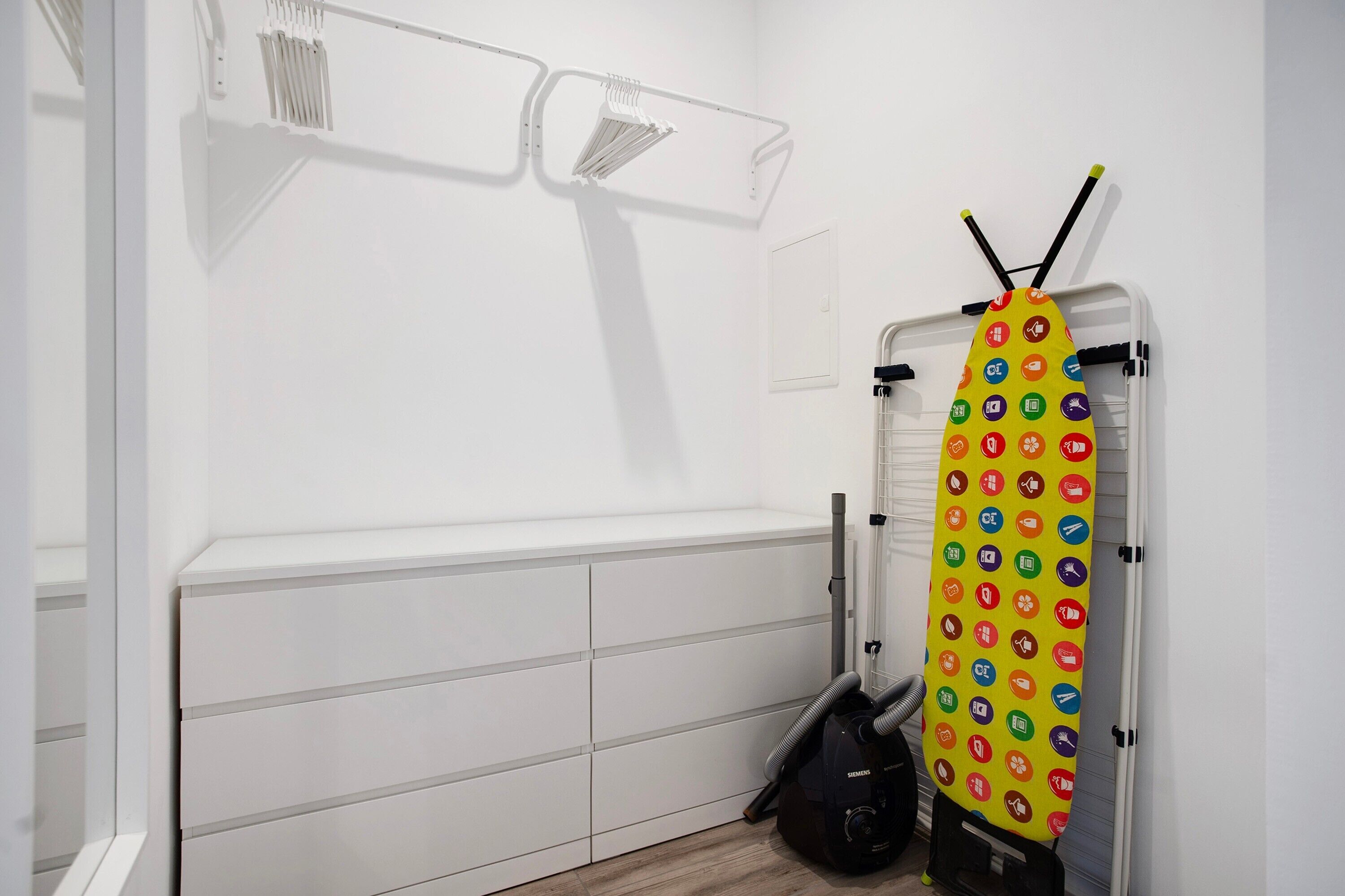 Una habitación con un armario blanco, un aspirador y una tabla de planchar colorida.