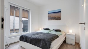 Un dormitorio con cama, mesitas de noche, un cuadro y vistas a través de una ventana.