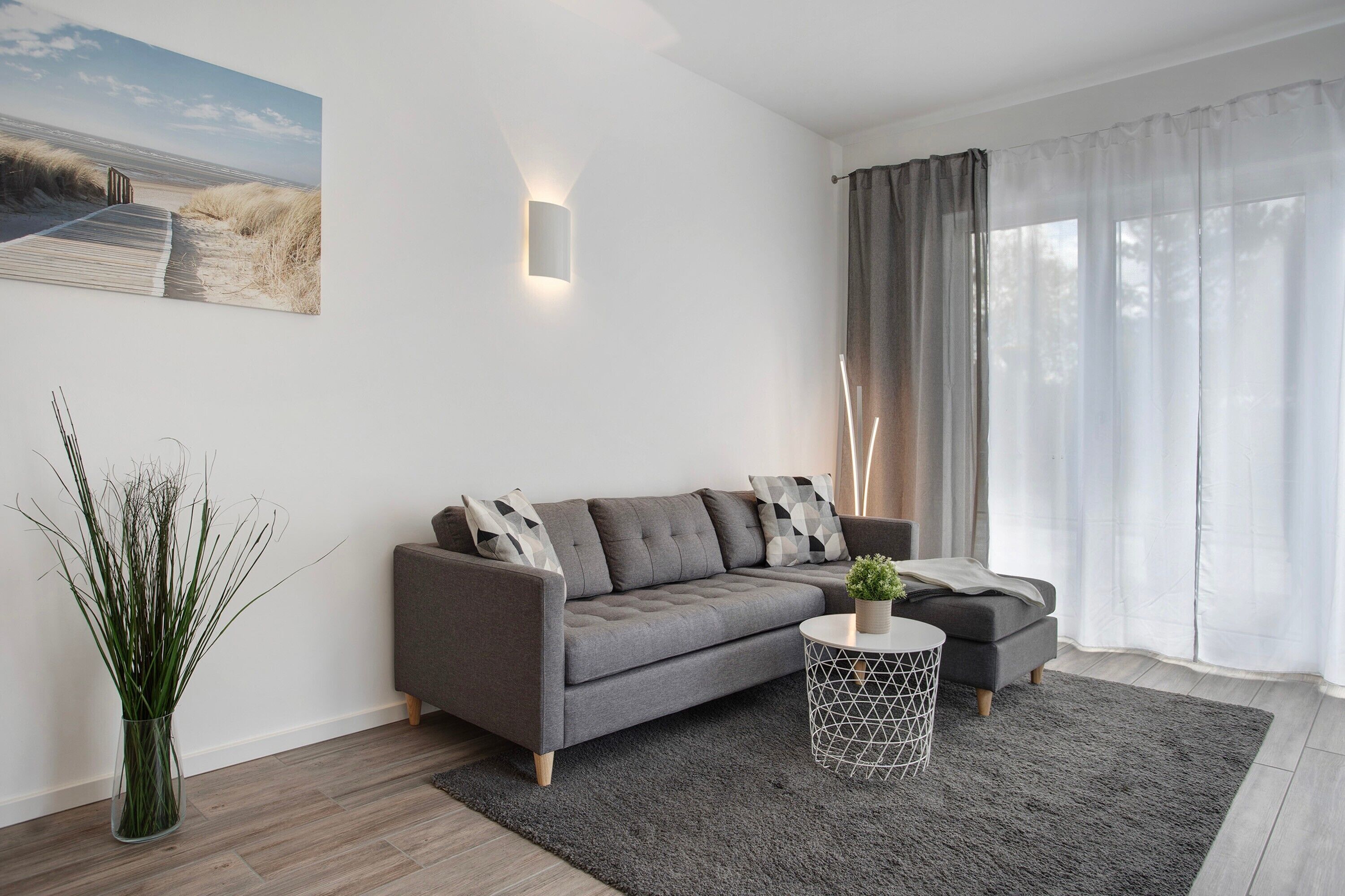 Ein modernes Wohnzimmer mit grauem Sofa, einem kleinen Beistelltisch, einer Pflanzvase und einem Bild an der Wand.