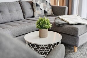A grey sofa with a geometric pillow, a round table with a woven base, and a small potted plant.