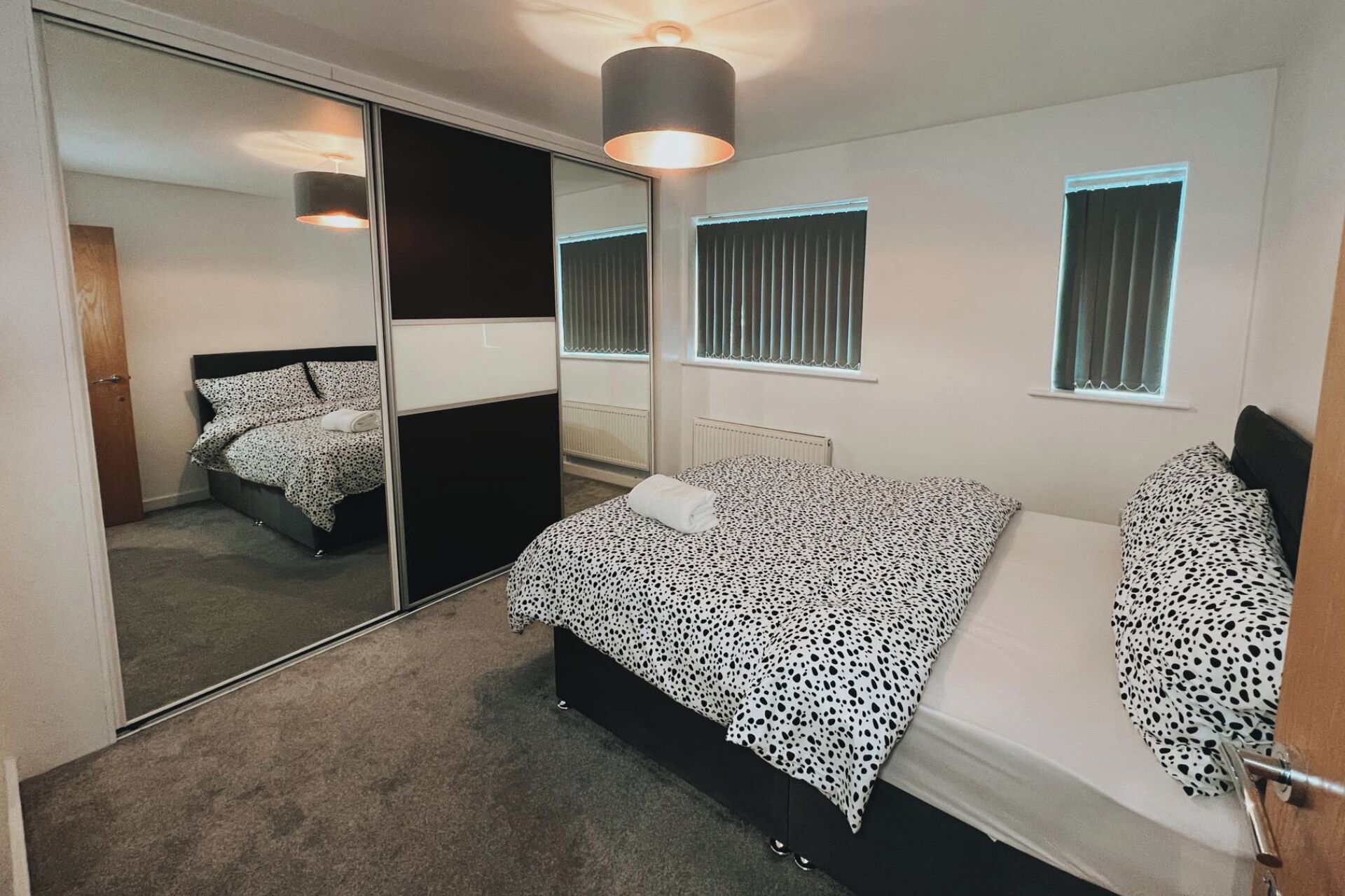A bedroom with a bed, a mirror, and two windows with blinds.
