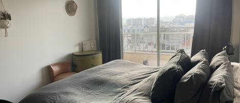 A bedroom with a large bed, a nightstand, a radiator, and a view of the city through a window.