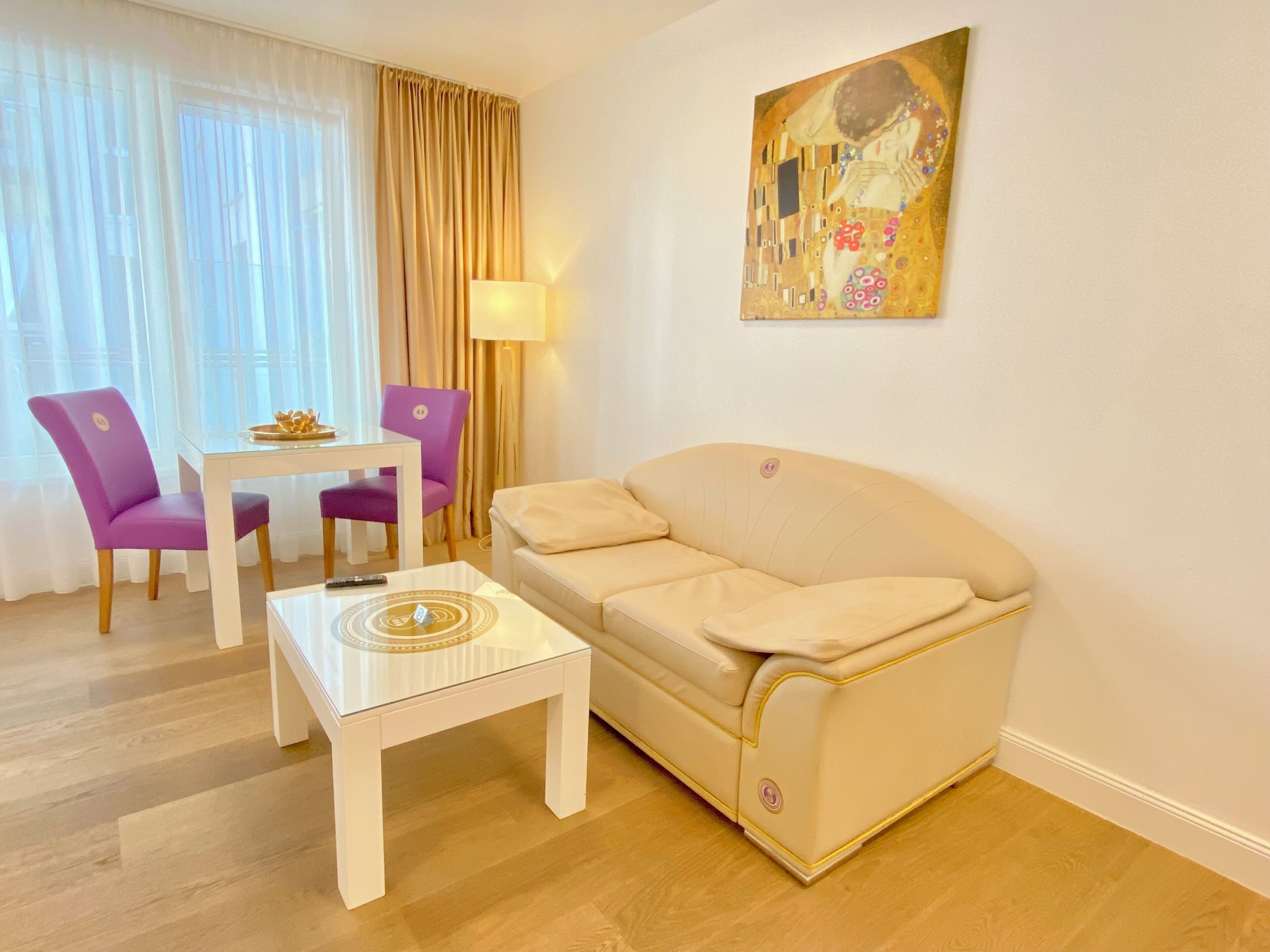 Una sala de estar con un sofá beige, una mesa de centro de vidrio y dos sillas moradas. Hay un cuadro en la pared y una lámpara sobre la mesa.