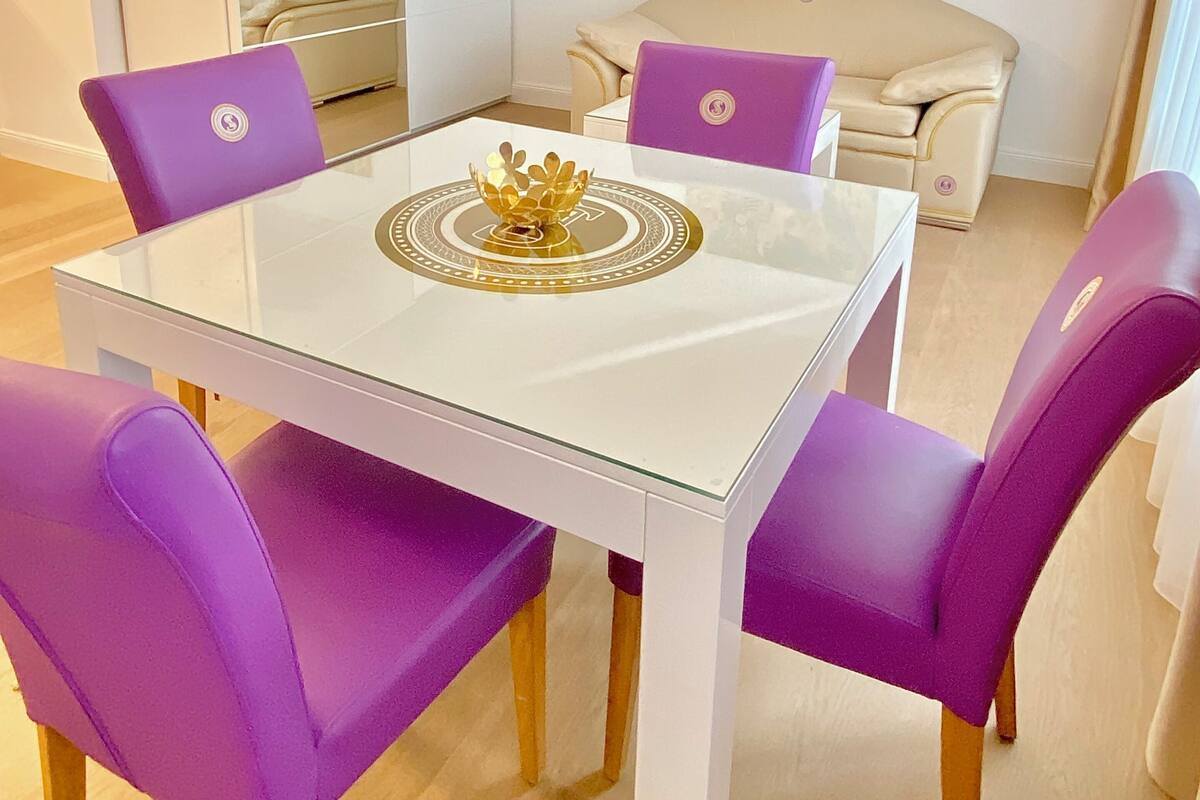 A dining area with a white table, purple chairs, a decorative golden crown centerpiece, a sofa, and a painting on the wall.