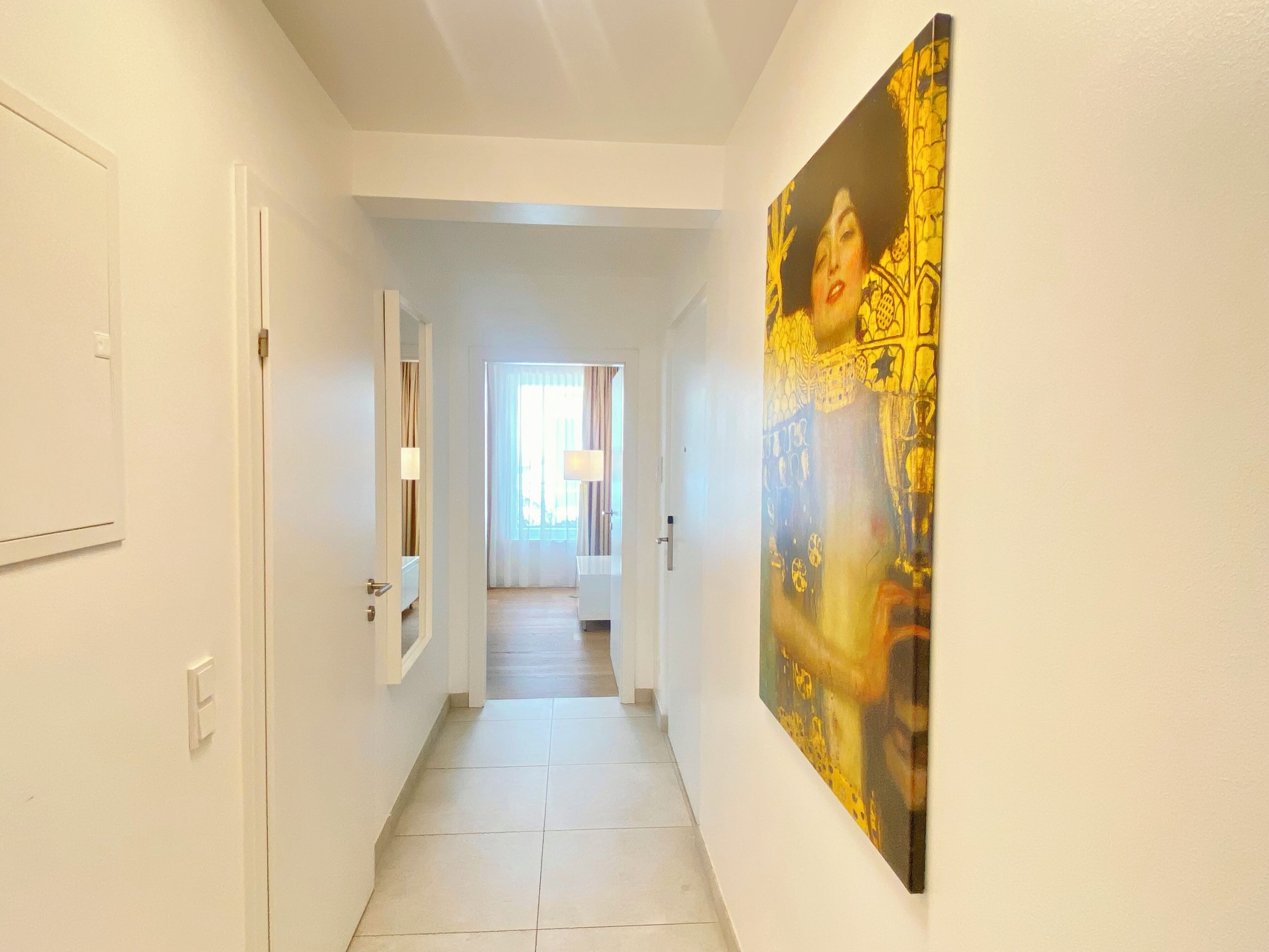 A hotel hallway with white walls, a tiled floor, and a large painting on the right wall.