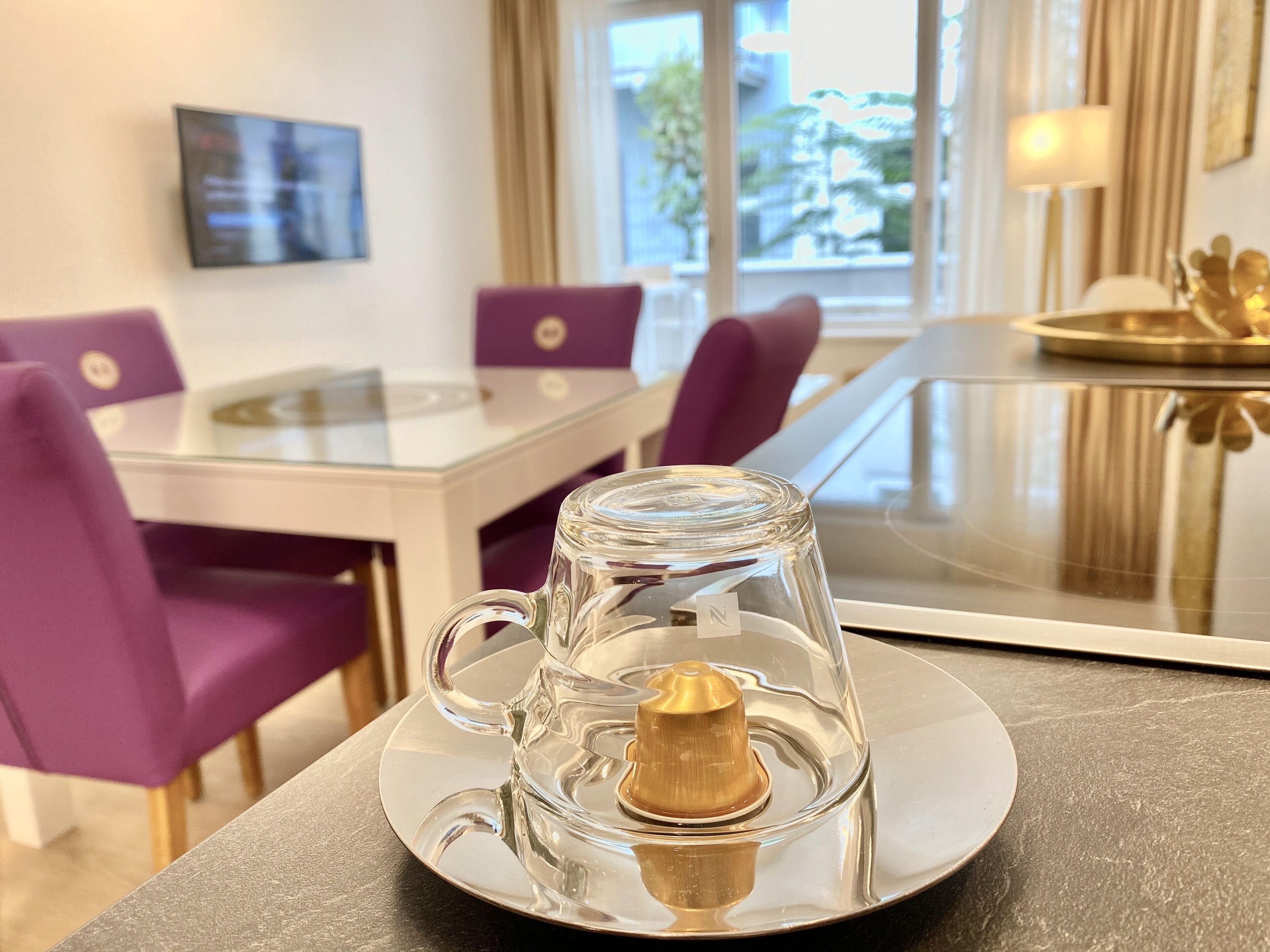 A clear glass mug with a gold lid on a reflective surface, a glass table, purple chairs, and a view of the outdoors through a window.