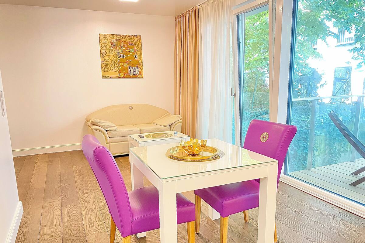 A dining area with a glass table, two purple chairs, a beige sofa, and a window with a view of greenery.