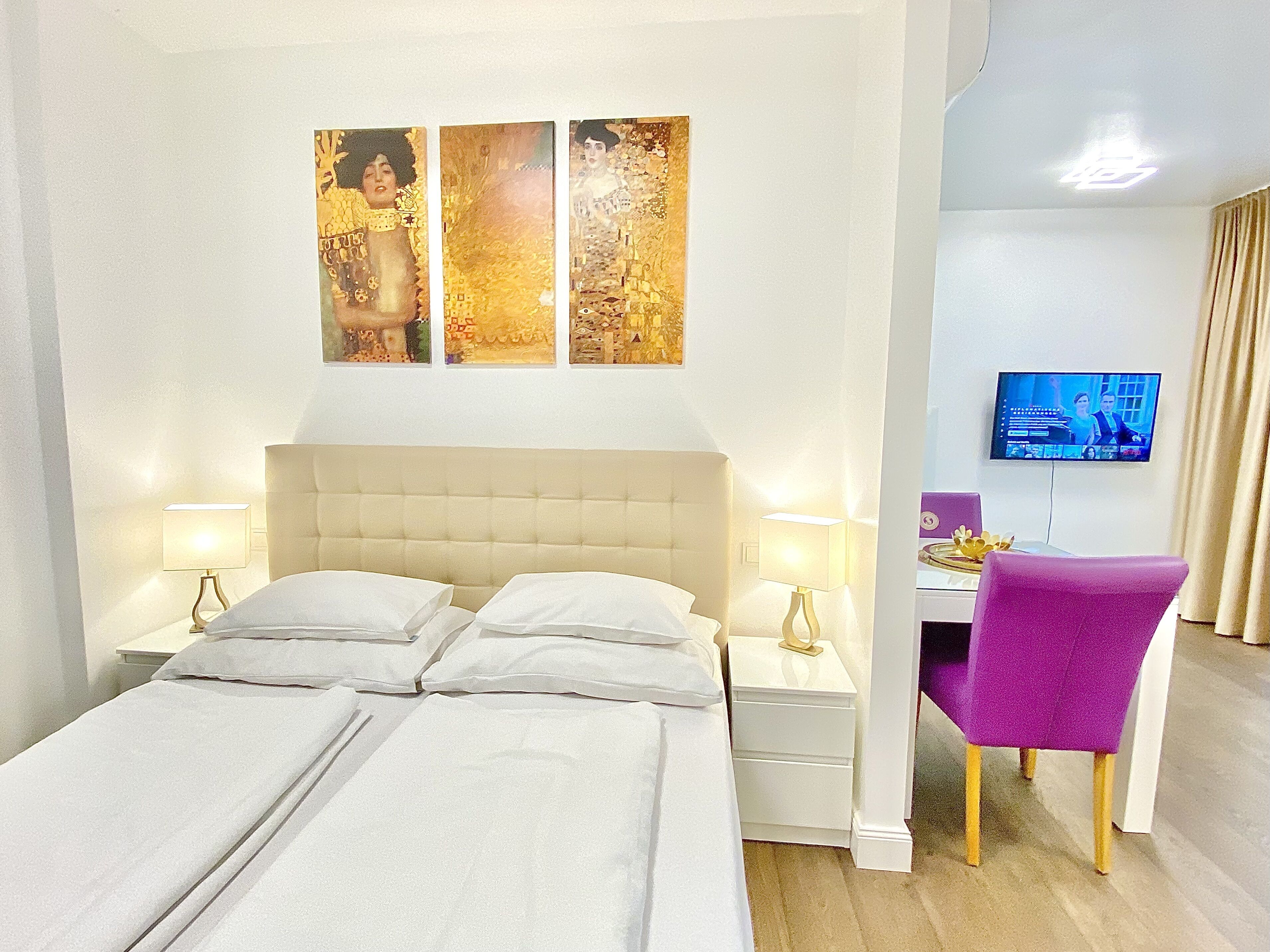 A hotel room with a bed, two bedside tables, a desk with a chair, a flat-screen TV, and three wall-mounted artworks.