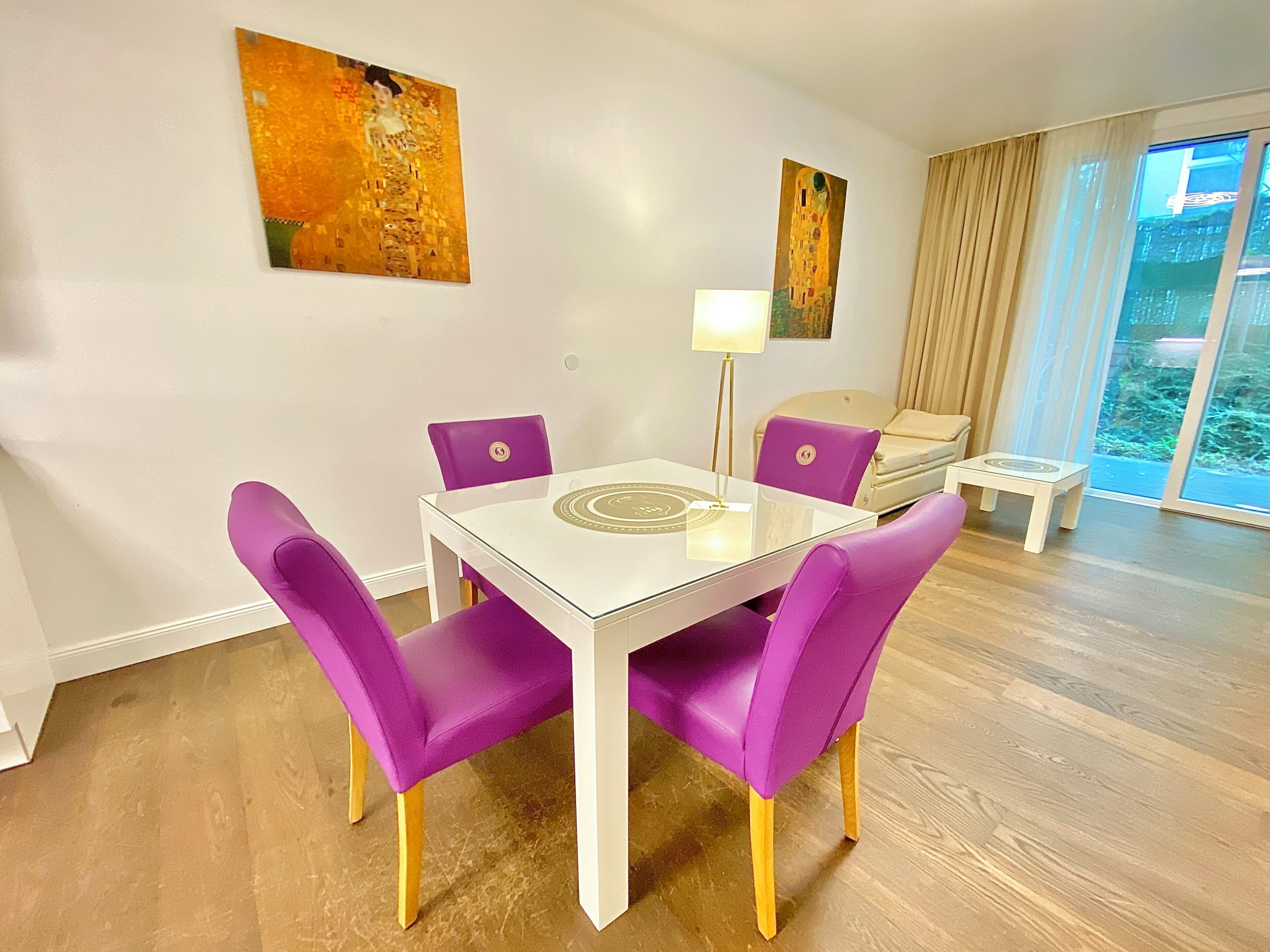 Een moderne woonkamer met een glazen eettafel, paarse stoelen, een bank, een klein bijzettafeltje en twee schilderijen aan de muur.