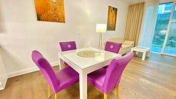 Een moderne woonkamer met een glazen eettafel, paarse stoelen, een bank, een klein bijzettafeltje en twee schilderijen aan de muur.