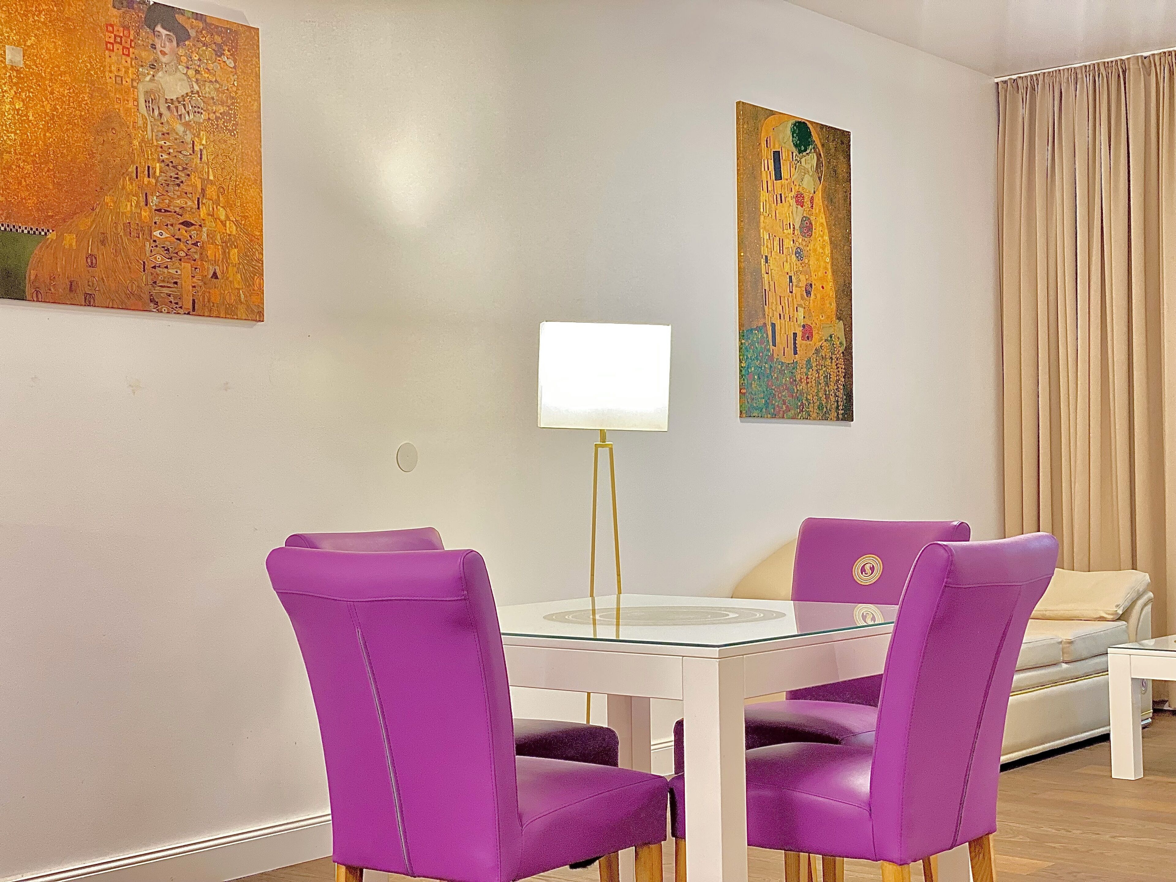 A modern dining area with a glass table and purple chairs, a white lamp, and two paintings on the wall.