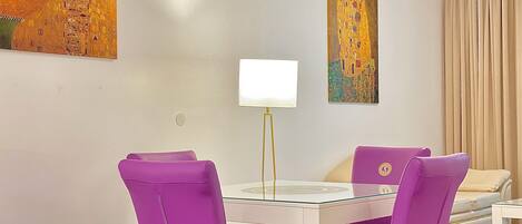 A modern dining area with a glass table and purple chairs, a white lamp, and two paintings on the wall.
