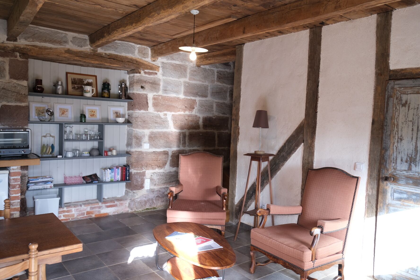 A rustic interior with stone walls, wooden beams, a fireplace, and a cozy seating area with two chairs and a table.