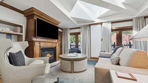 A modern living room with a fireplace, comfortable seating, a flat-screen TV, and large windows offering a view of the outside.