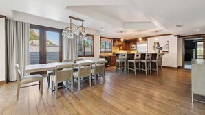 A spacious kitchen with wooden flooring, a dining area with a large table and chairs, and a view of the outdoors through large windows.