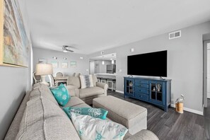 A modern living room with a sectional sofa, a flat-screen TV, and a dining area in the background. - 6 Beds, Pool, Corner View- 3 Bdrm Duneside Dream (North Myrtle Beach)