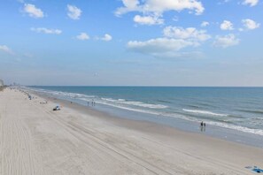 A wide sandy beach with gentle waves, a clear sky, and a few people in the distance. - 6 Beds, Pool, Corner View- 3 Bdrm Duneside Dream (North Myrtle Beach)