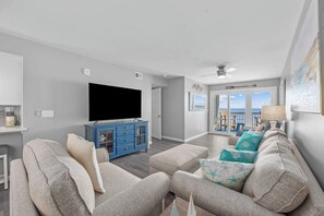 A living room with a flat-screen TV, a blue cabinet, a grey sofa set, and a view to the outside. - 6 Beds, Pool, Corner View- 3 Bdrm Duneside Dream (North Myrtle Beach)