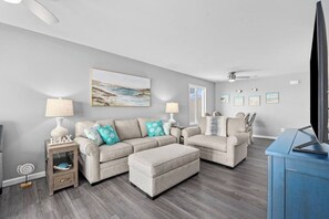 A modern living room with a grey sofa set, a coffee table, a TV stand, and a ceiling fan. - 6 Beds, Pool, Corner View- 3 Bdrm Duneside Dream (North Myrtle Beach)