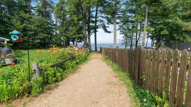 Un chemin de gravier bordé d’une clôture en bois et d’une végétation luxuriante, qui mène à un plan d’eau.