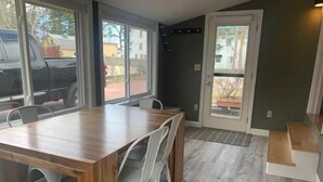 Un espace repas avec une table en bois et des chaises blanches, de grandes fenêtres et une porte donnant sur l’extérieur.