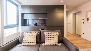 A modern living room with a grey sofa and two patterned throw pillows, a kitchen with dark cabinets and a marble backsplash, and a hallway with wooden flooring and a framed picture on the wall. - RUAH Q - Quiet Flat by LovelyStay (Vila Nova de Gaia)