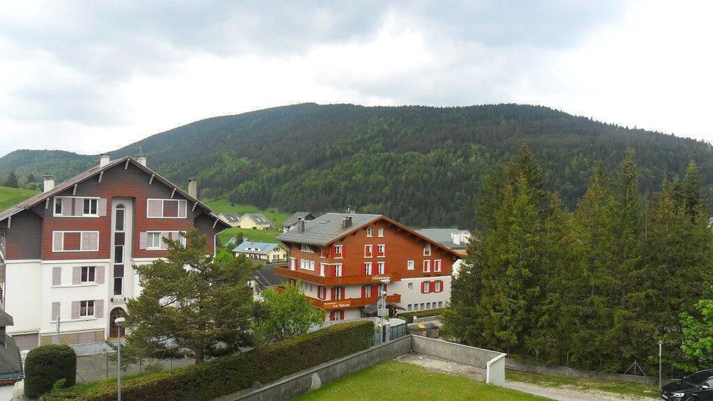 A multi-story building with a red facade and white trim, surrounded by lush greenery and a forested mountain in the background.
