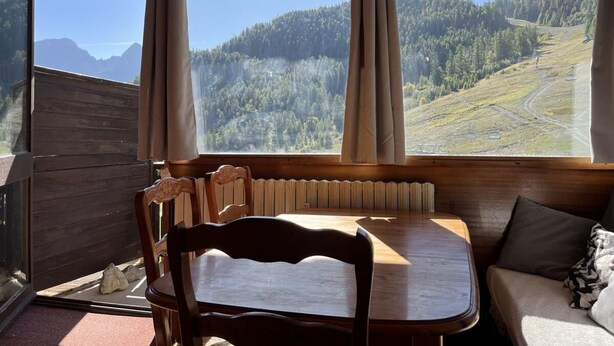 A cozy cabin interior with a wooden table and chairs, a sofa, and a large window offering a view of a forested mountain landscape. - 200 meters from the ski slopes, 3rd floor, balcony, TV, ski locker, 50 m², Pra Loup (Uvernet-Fours)