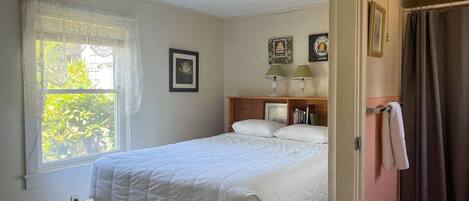 A bedroom with a bed, a window with curtains, a bedside table with lamps, and a door to a bathroom.