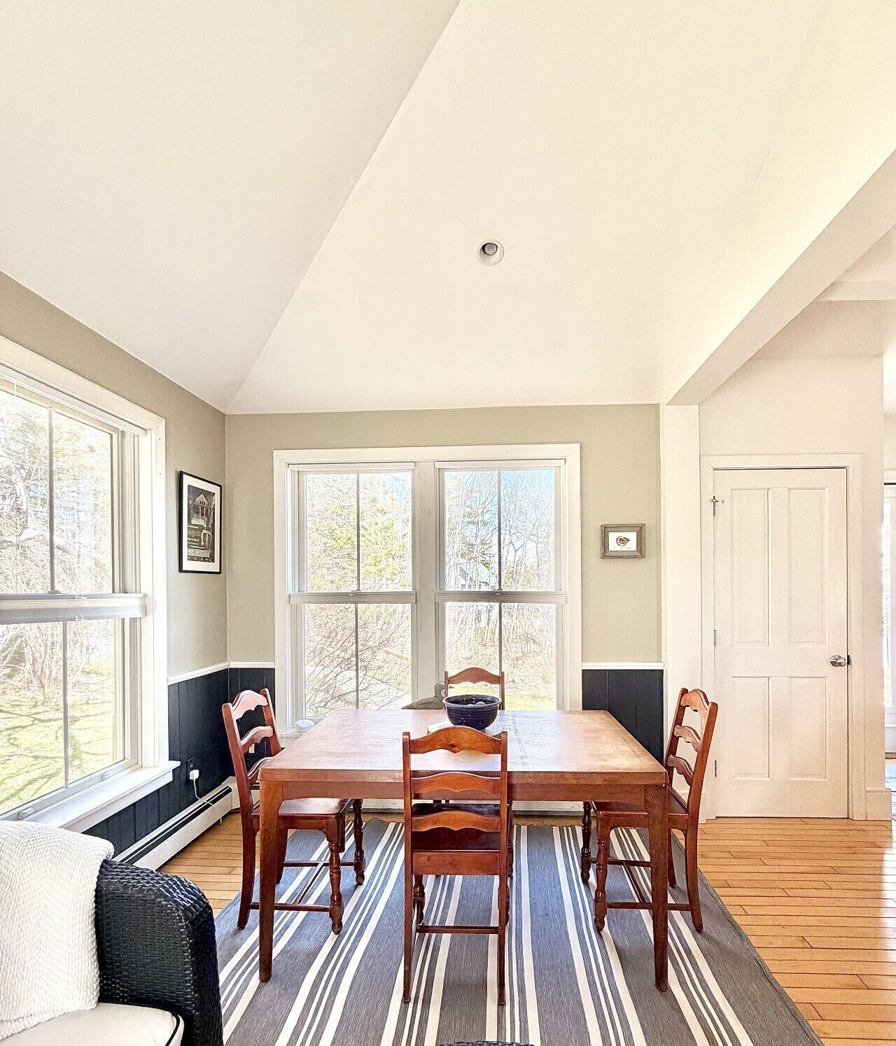 A dining room with a wooden table, four chairs, a bowl, and a window with a view of trees.