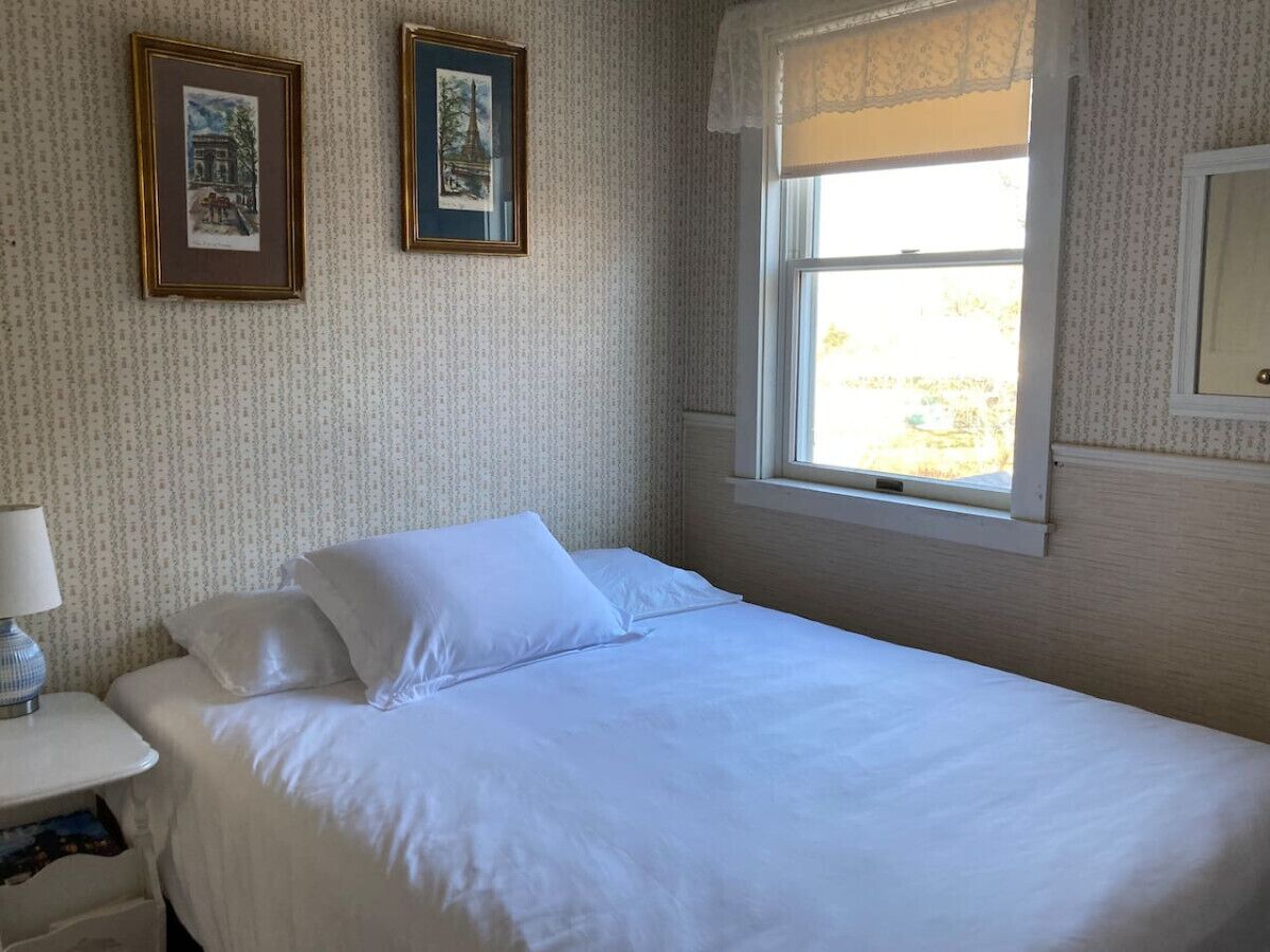 A neatly made bed in a room with patterned wallpaper, a window with a white blind, and two framed pictures on the wall.