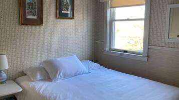 A neatly made bed in a room with patterned wallpaper, a window with a white blind, and two framed pictures on the wall.