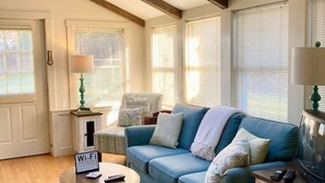 A cozy living room with a blue sofa, a coffee table, and a TV stand. There is a ceiling fan, a skylight, and a door with a Wi-Fi sign. - Orchid Harbor, Bailey Island 3BR (Harpswell)