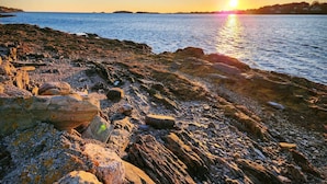 A rocky shoreline at sunset with calm water and distant land. - Orchid Harbor, Bailey Island 3BR (Harpswell)