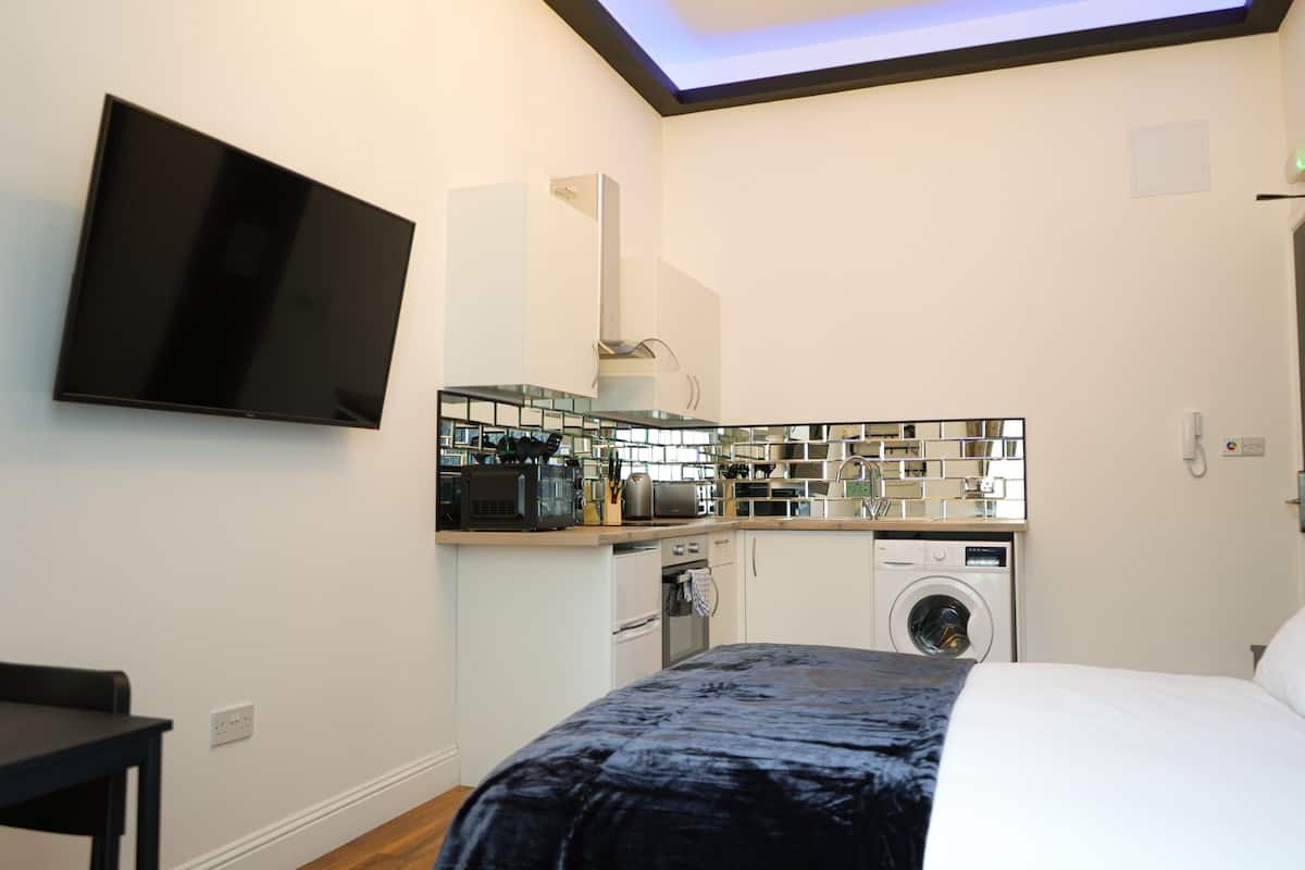 Habitación de hotel compacta con cama, televisor de pantalla plana montado en la pared, kitchenette con microondas, tostadora y lavadora, además de una pequeña mesa.