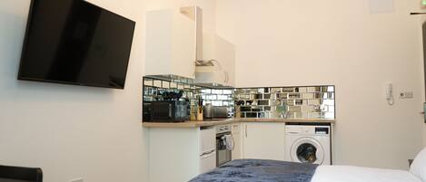 A compact hotel room with a bed, a flat-screen TV mounted on the wall, a kitchenette with a microwave, toaster, and washing machine, and a small table.