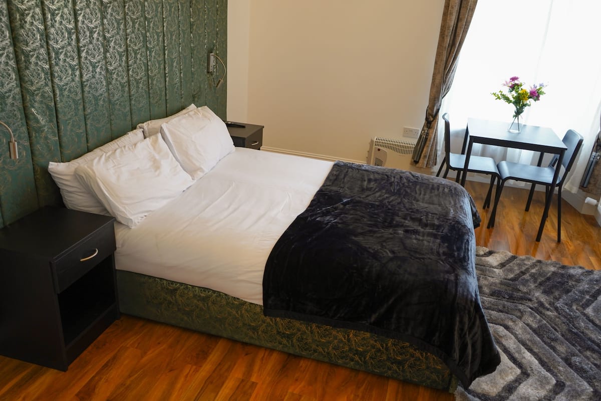 Un dormitorio con una cama, una mesita de noche, una mesa con sillas y una ventana con cortinas.