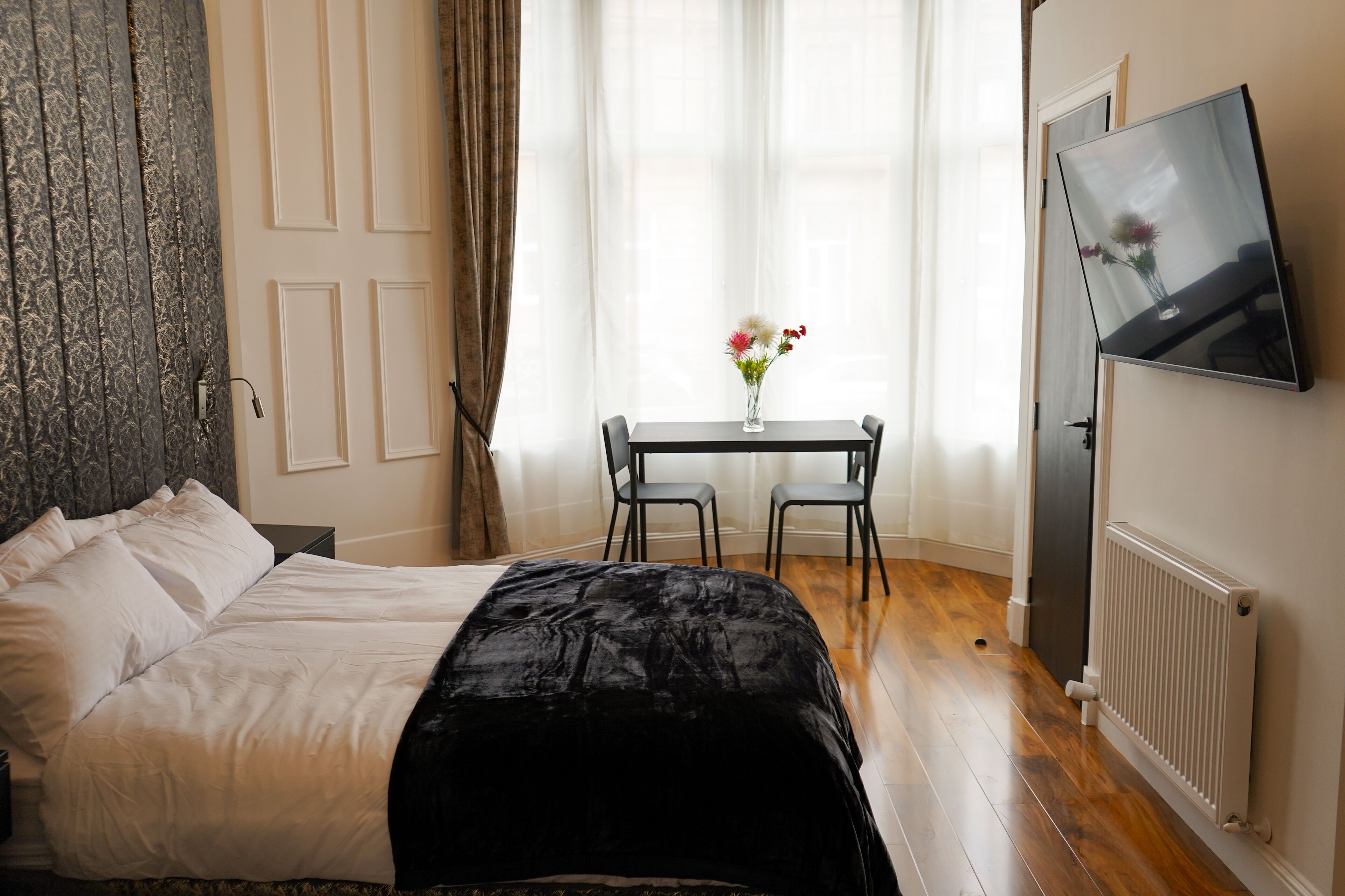 A hotel room with a bed, a small table with a vase of flowers, a TV mounted on the wall, and a window with curtains.