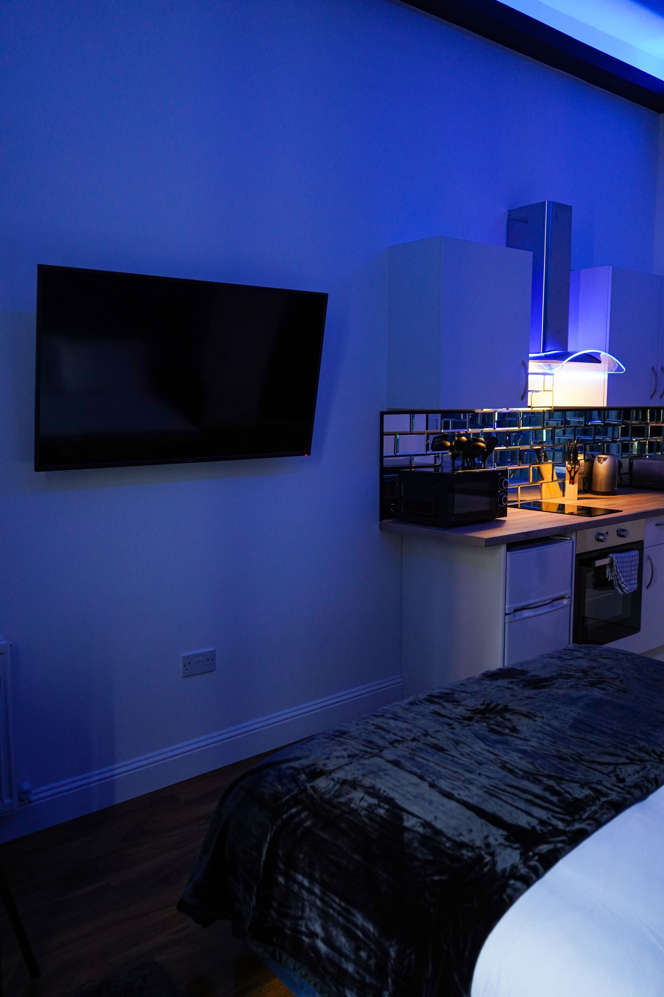 A modern bedroom with a bed, a flat-screen TV mounted on the wall, a kitchenette with a microwave, toaster, and kettle, and a small oven.