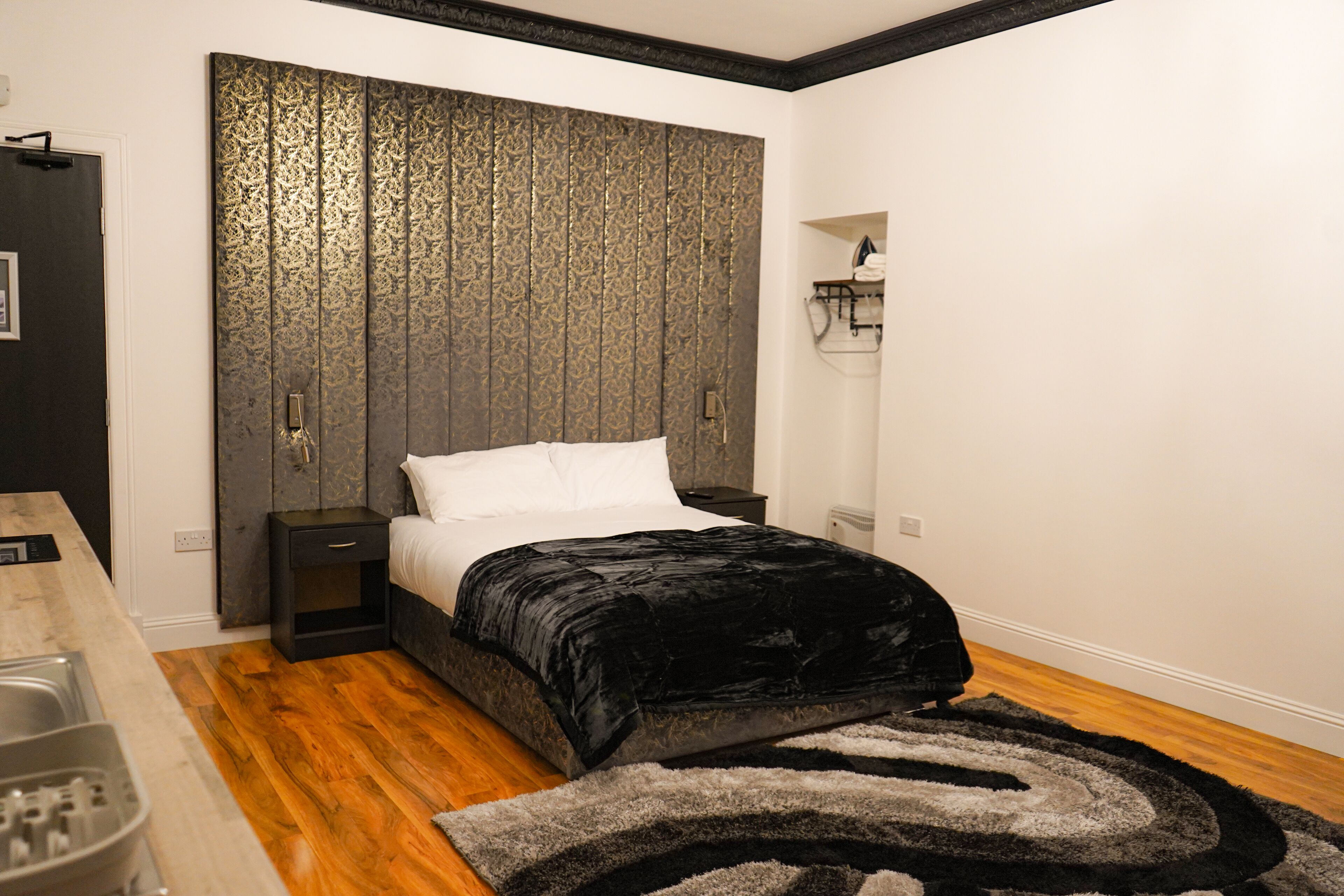 A bedroom with a bed, a nightstand, a dresser, a rug, and a wall-mounted light fixture.