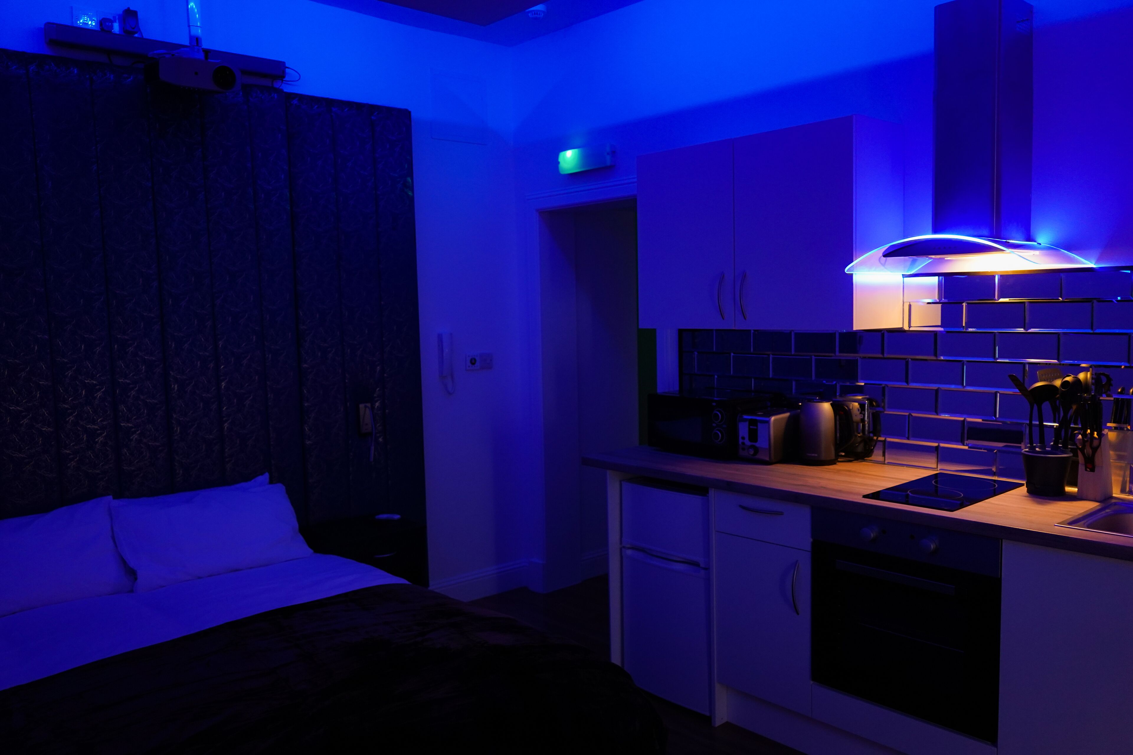 A modern kitchen with a built-in oven, microwave, and coffee maker. There is a bed with white bedding in the adjacent room.