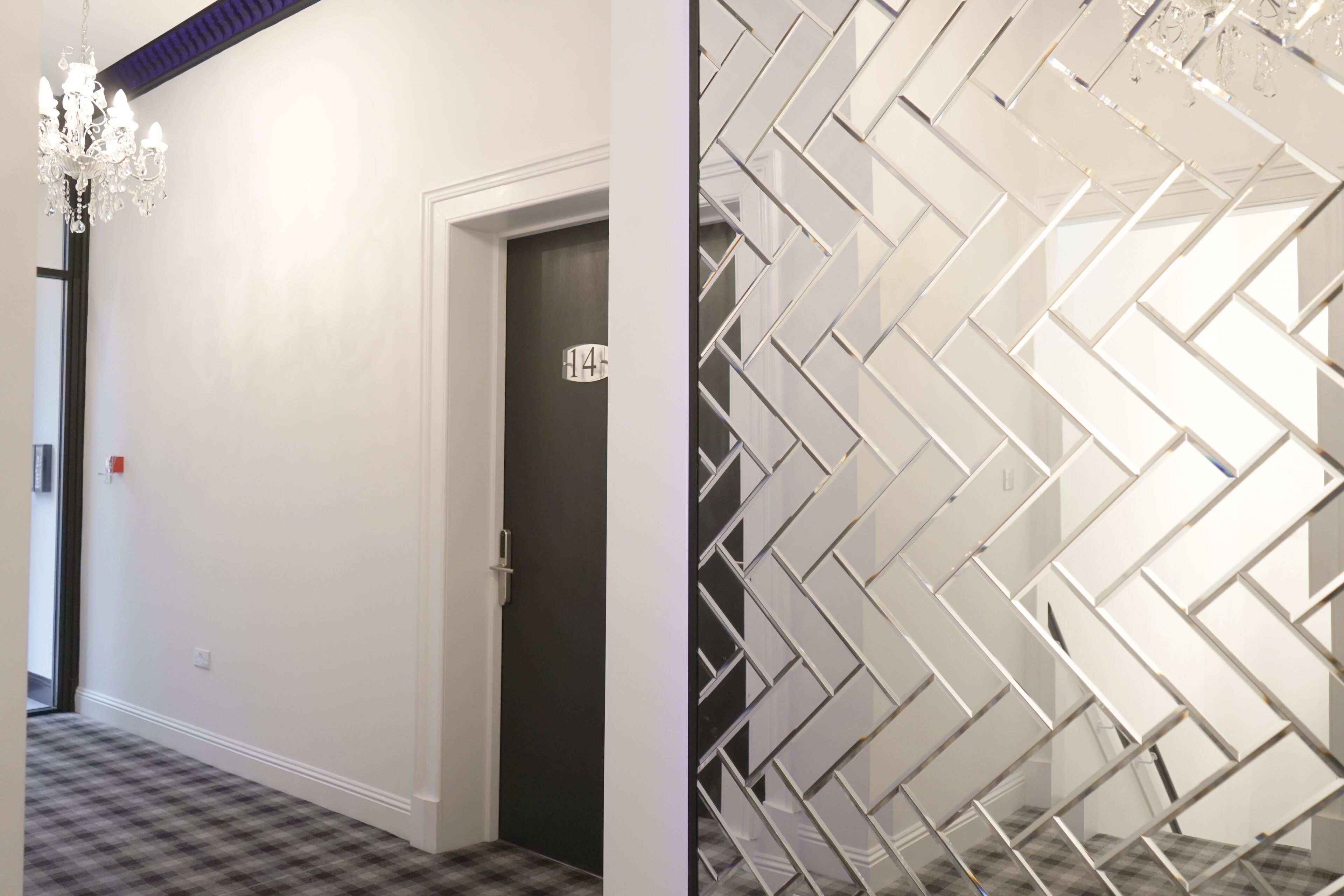 Een gang met een geruit tapijt, een kroonluchter, een deur met een bordje en een muur met een geometrisch patroon.