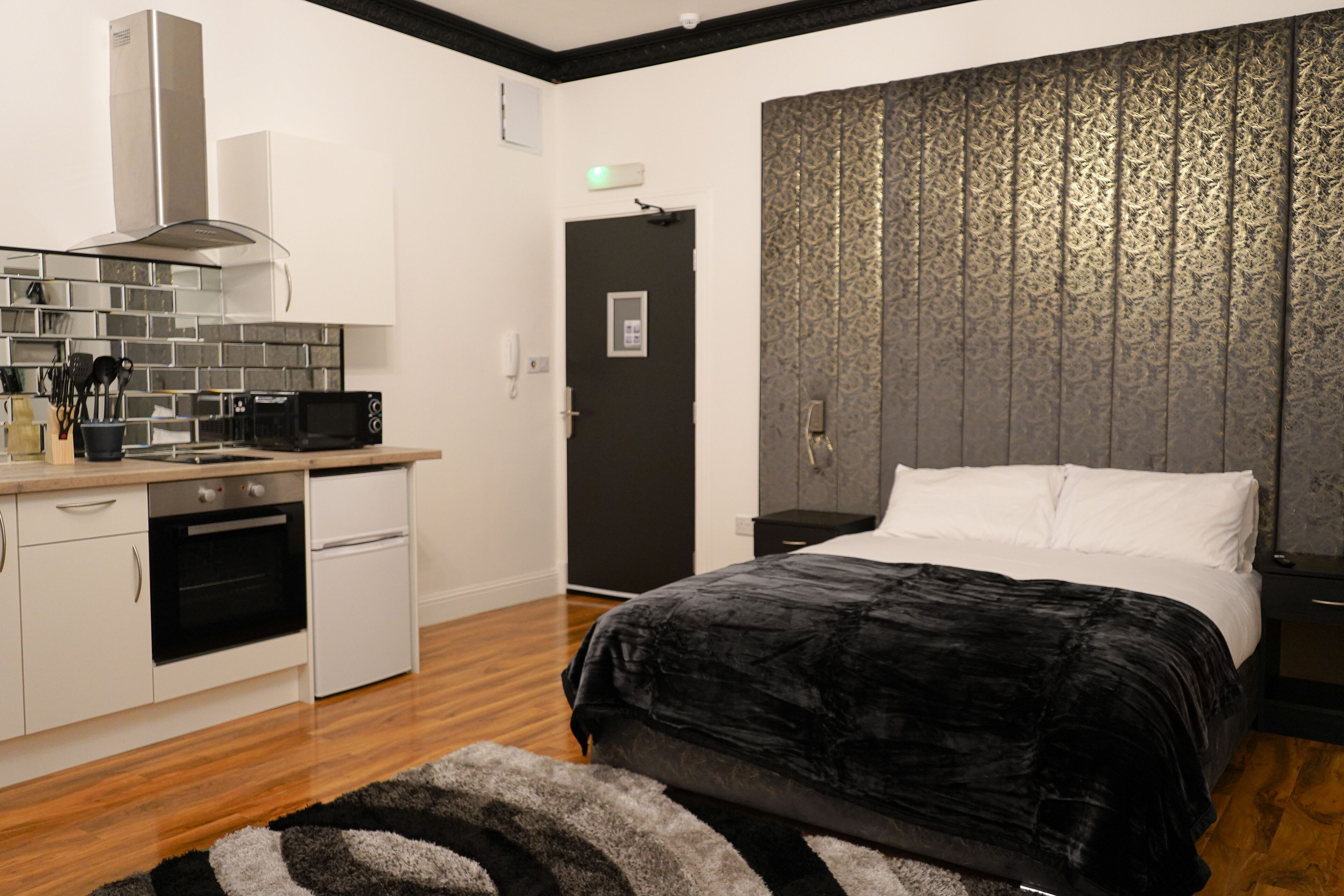 Habitación de hotel pequeña con kitchenette, cama con colcha negra y papel tapiz estampado en la pared.
