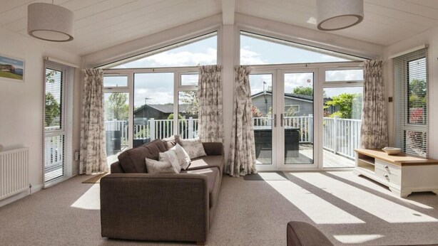 A spacious living room with a large sofa, a coffee table, and a view of a fenced backyard through large sliding glass doors. - Lodge Sleeps 6 With Private Hot Tub (Honiton)