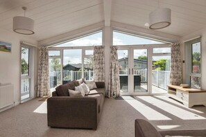 A spacious living room with a large sofa, a coffee table, and a view of a fenced backyard through large sliding glass doors. - Lodge Sleeps 6 With Private Hot Tub (Honiton)