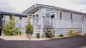A mobile home with a covered porch, white exterior, and a fenced yard with small plants. - Lodge Sleeps 6 With Private Hot Tub (Honiton)
